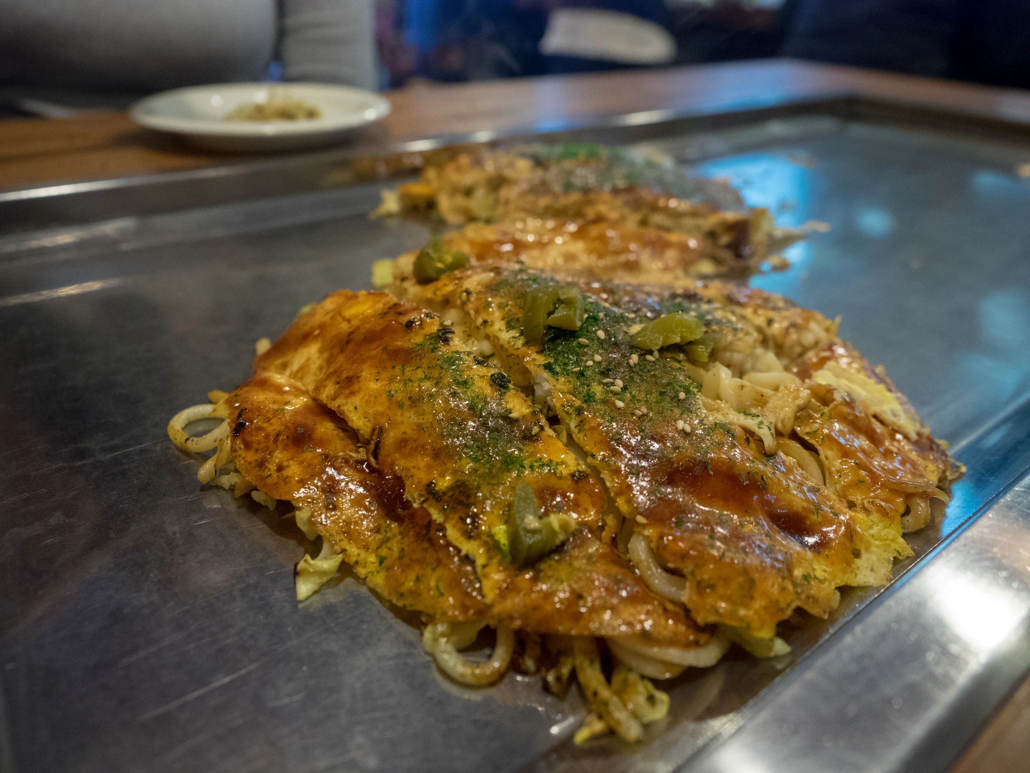Freshly assembled Hiroshima-style okonomiyaki with shredded mountain cabbage, pork belly, and savory Otafuku sauce on hot teppan