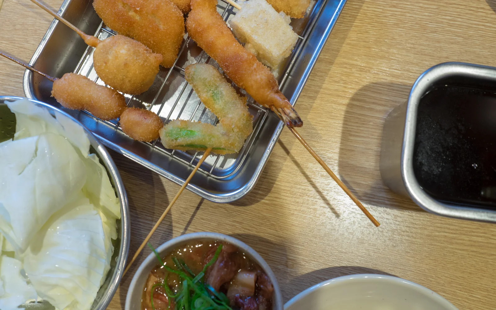 Golden panko-crusted deep fried kushikatsu skewers resting in a stainless steel tray with raw cabbage at Shinsekai