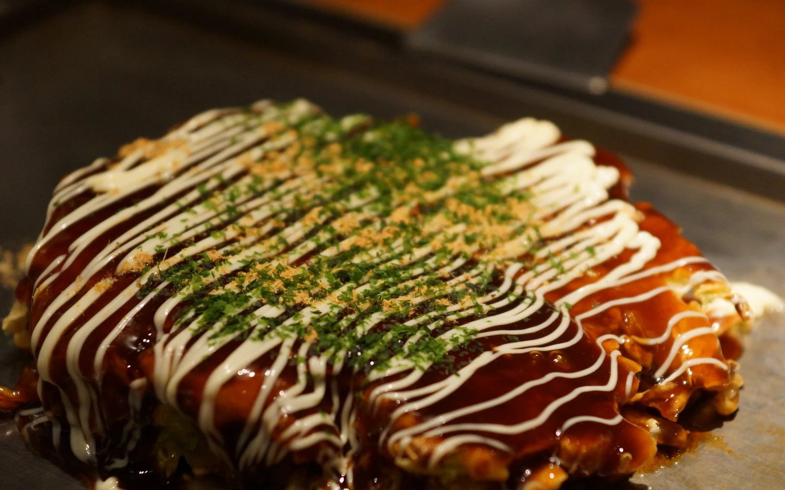 Thick cabbage-loaded okonomiyaki sizzling on a flat-top teppan with sliced pork belly and savory glaze