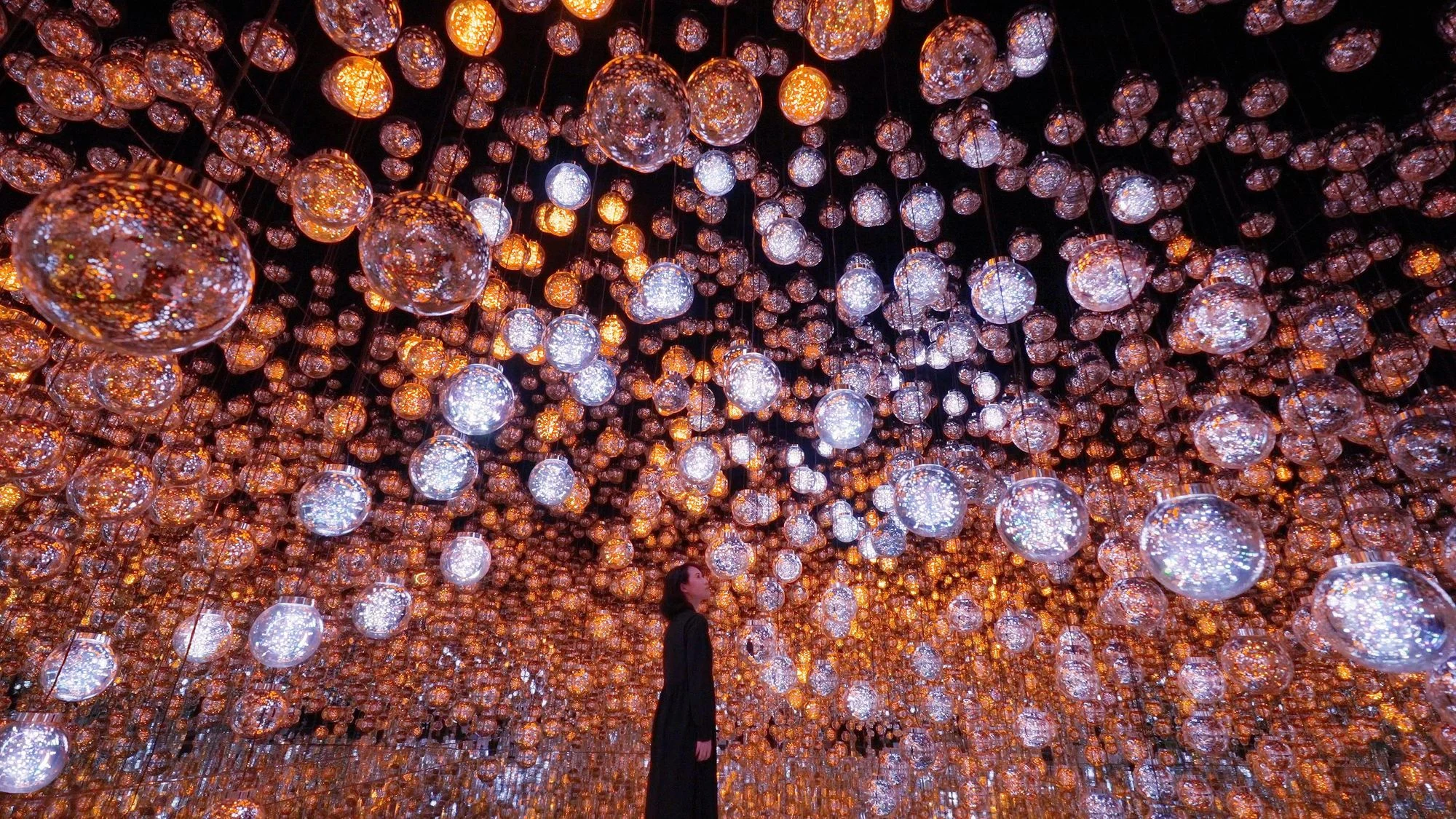 A visitor stands among glowing suspended spheres and reflections in Bubble Universe at teamLab Borderless.